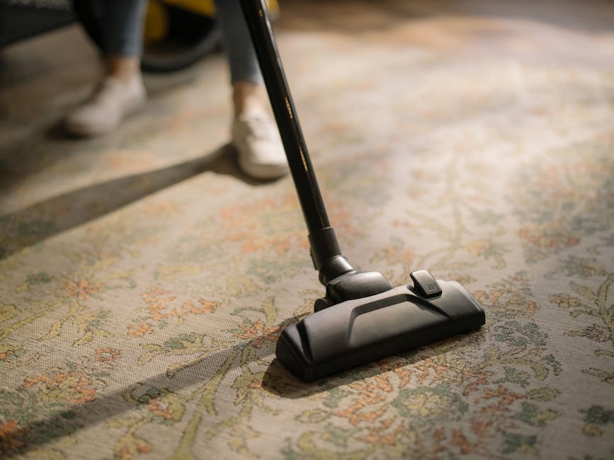 A woman with curly hair wearing a beige top, dark jeans, and white shoes is performing surface cleaning in a living room. She is using a yellow vacuum cleaner with a black hose attachment on a pattern