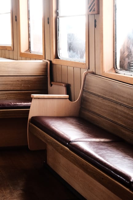 Interior view of a wooden seating area with built-in benches along a wall, featuring polished dark brown leather cushions. The benches are situated under large, clear glass windows with wooden frames, allowing natural light to illuminate the space. The wooden surfaces are smooth and well-maintained, indicating a high standard of cleaning and upkeep, typical of a space requiring surface cleaning and sanitisation. Visible tools or cleaning products are not present, but the area appears neat and hygienic, consistent with professional domestic cleaning practices, as promoted by Cleaners Greenwich in their upholstery cleaning and stain removal tips for the Cutty Sark area.