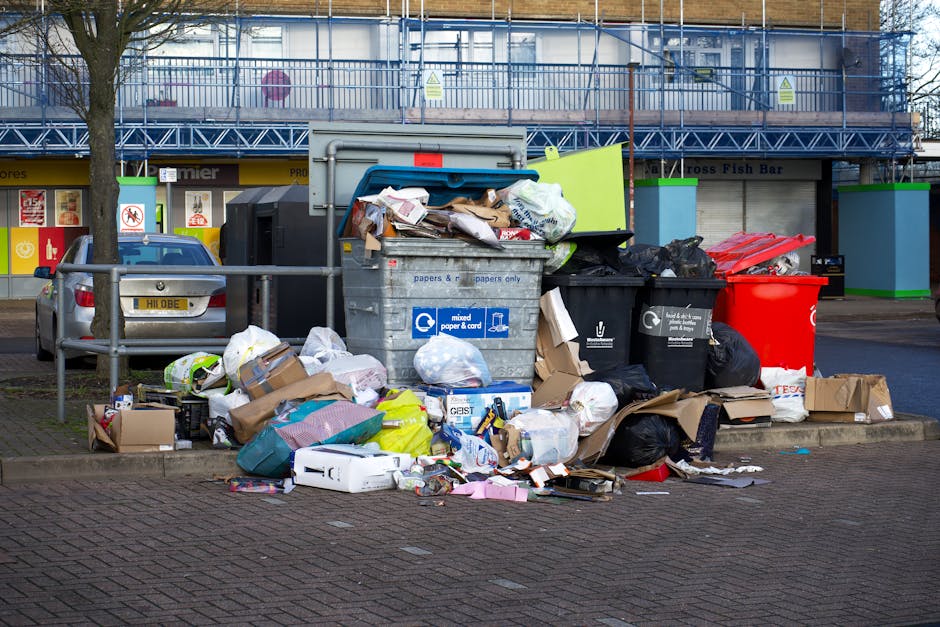 A large outdoor waste collection area with multiple overflowing recycling and general waste bins, including a grey mixed paper and card bin, a black general waste bin, and a red recycling bin. The ground around the bins is littered with various discarded cardboard boxes, paper, plastic bags, and packaging materials, some spilling onto the paved surface. The background features a building with a blue metal facade and a storefront on the ground level, with signage indicating local businesses. The scene is outdoors during daylight with natural lighting, and the waste containers are positioned on a paved sidewalk area, partially enclosed with safety railings. This image illustrates improper waste disposal and highlights the importance of adhering to waste and cleaning disposal regulations, as detailed by Cleaners Greenwich on their website.