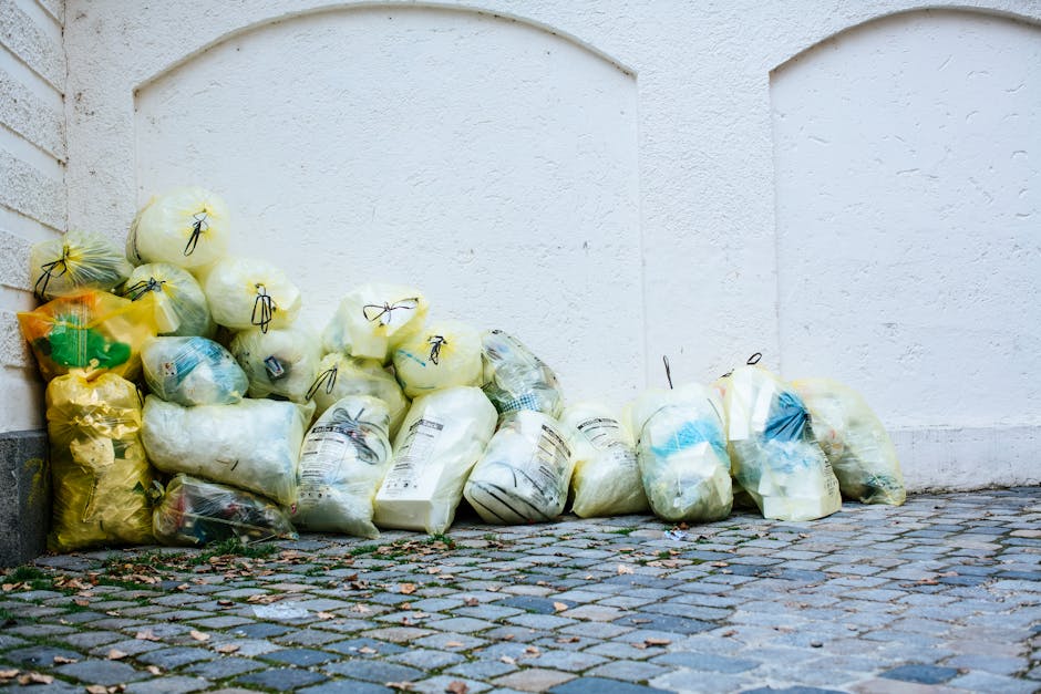 A pile of yellow, white, and transparent rubbish bags filled with waste is stacked against a white wall outside a building. The bags are made of plastic, some with printed labels, and appear to contain household waste. The surface beneath the bags is a cobblestone pavement with scattered leaves and small debris. The scene is well-lit with natural daylight, highlighting the cleanliness and orderliness of the waste collection area. This image relates to surface cleaning and waste disposal, reflecting hygienic waste management practices consistent with Greenwich Council rules for waste and cleaning disposal, as promoted by Cleaners Greenwich.