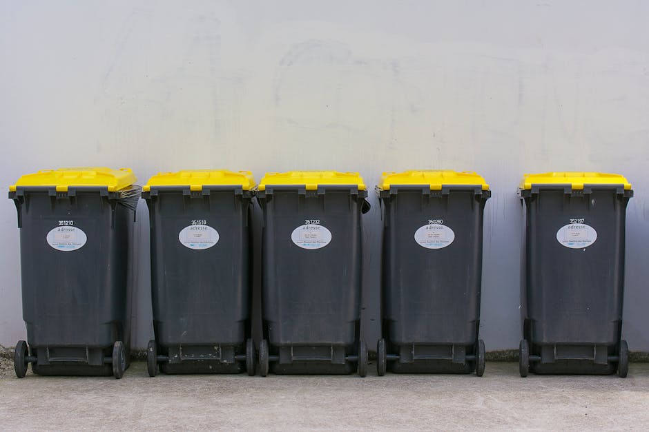 A large outdoor waste collection area with multiple overflowing recycling and general waste bins, including a grey mixed paper and card bin, a black general waste bin, and a red recycling bin. The ground around the bins is littered with various discarded cardboard boxes, paper, plastic bags, and packaging materials, some spilling onto the paved surface. The background features a building with a blue metal facade and a storefront on the ground level, with signage indicating local businesses. The scene is outdoors during daylight with natural lighting, and the waste containers are positioned on a paved sidewalk area, partially enclosed with safety railings. This image illustrates improper waste disposal and highlights the importance of adhering to waste and cleaning disposal regulations, as detailed by Cleaners Greenwich on their website.