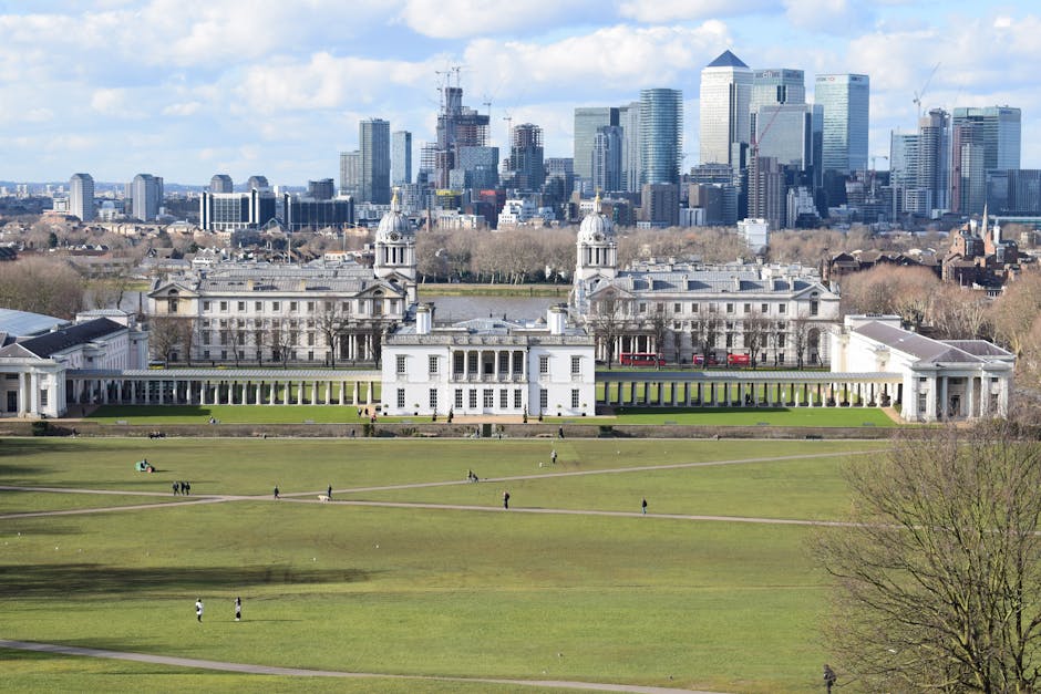 A wide view of a historic white building with classical architecture, including symmetrical wings and a central section topped with domed towers, situated in a large, well-maintained grassy park with walking paths and a few visitors. In the background, a modern city skyline with tall skyscrapers and construction cranes is visible under a partly cloudy sky. The scene reflects an urban setting combining heritage architecture and contemporary development, with lush green lawns and a clear atmosphere. The image is related to residential or commercial cleaning services offered by Cleaners Greenwich, emphasizing the importance of maintaining hygiene and cleanliness in both historic and modern environments.