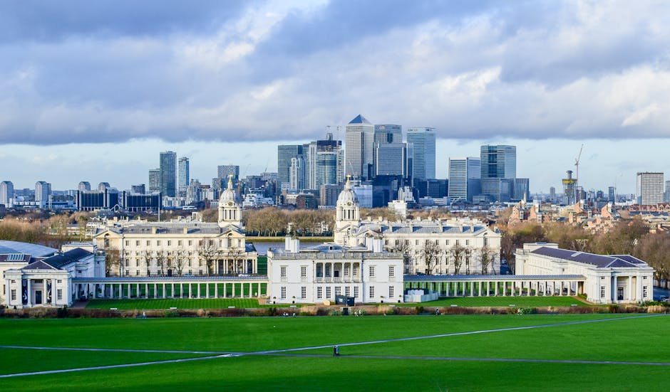 A panoramic view of a historic white building with classical architecture, featuring two prominent domed towers, set in well-maintained green lawns and bordered by a colonnade. In the background, a modern urban skyline with tall glass office buildings under a partly cloudy sky. The scene is bright with natural daylight, showcasing the clean and tidy appearance of the grounds. This image reflects property maintenance and the importance of surface cleaning and general hygiene, aligning with services offered by Cleaners Greenwich, especially for same-day house cleaning in Greenwich availability guide.