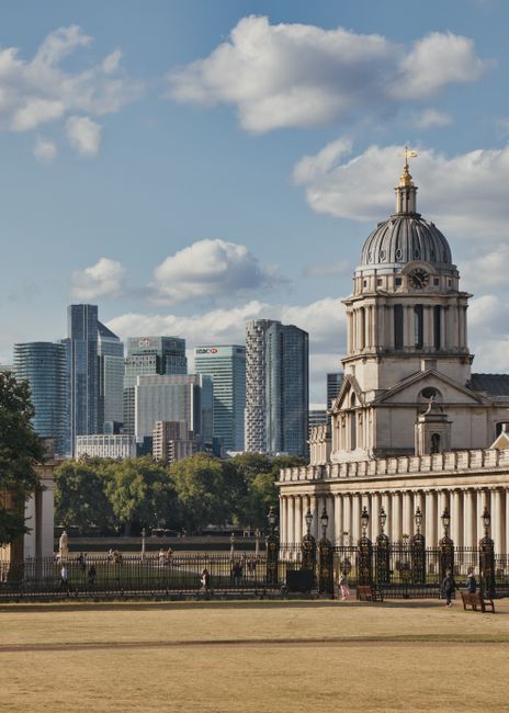 A wide view of a historic white building with classical architecture, including symmetrical wings and a central section topped with domed towers, situated in a large, well-maintained grassy park with walking paths and a few visitors. In the background, a modern city skyline with tall skyscrapers and construction cranes is visible under a partly cloudy sky. The scene reflects an urban setting combining heritage architecture and contemporary development, with lush green lawns and a clear atmosphere. The image is related to residential or commercial cleaning services offered by Cleaners Greenwich, emphasizing the importance of maintaining hygiene and cleanliness in both historic and modern environments.