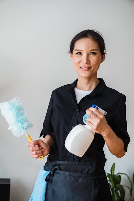 The image shows a female cleaner standing indoors with a plain light gray wall in the background. She is dressed in a black cleaning uniform with a white shirt underneath and has a blue apron tied around her waist. She is holding a white spray bottle in her right hand and a dusting tool with a blue and white fluffy attachment in her left hand. The surface of the area she is likely to clean includes polished wooden floors, and she appears ready to perform surface cleaning or sanitisation tasks. There are no other objects or clutter visible in the frame, emphasizing cleanliness and professional preparation. This image illustrates domestic cleaning services, such as surface decontamination or deep cleaning, consistent with the services offered by Cleaners Greenwich for end of tenancy cleaning costs and quotes in Greenwich.